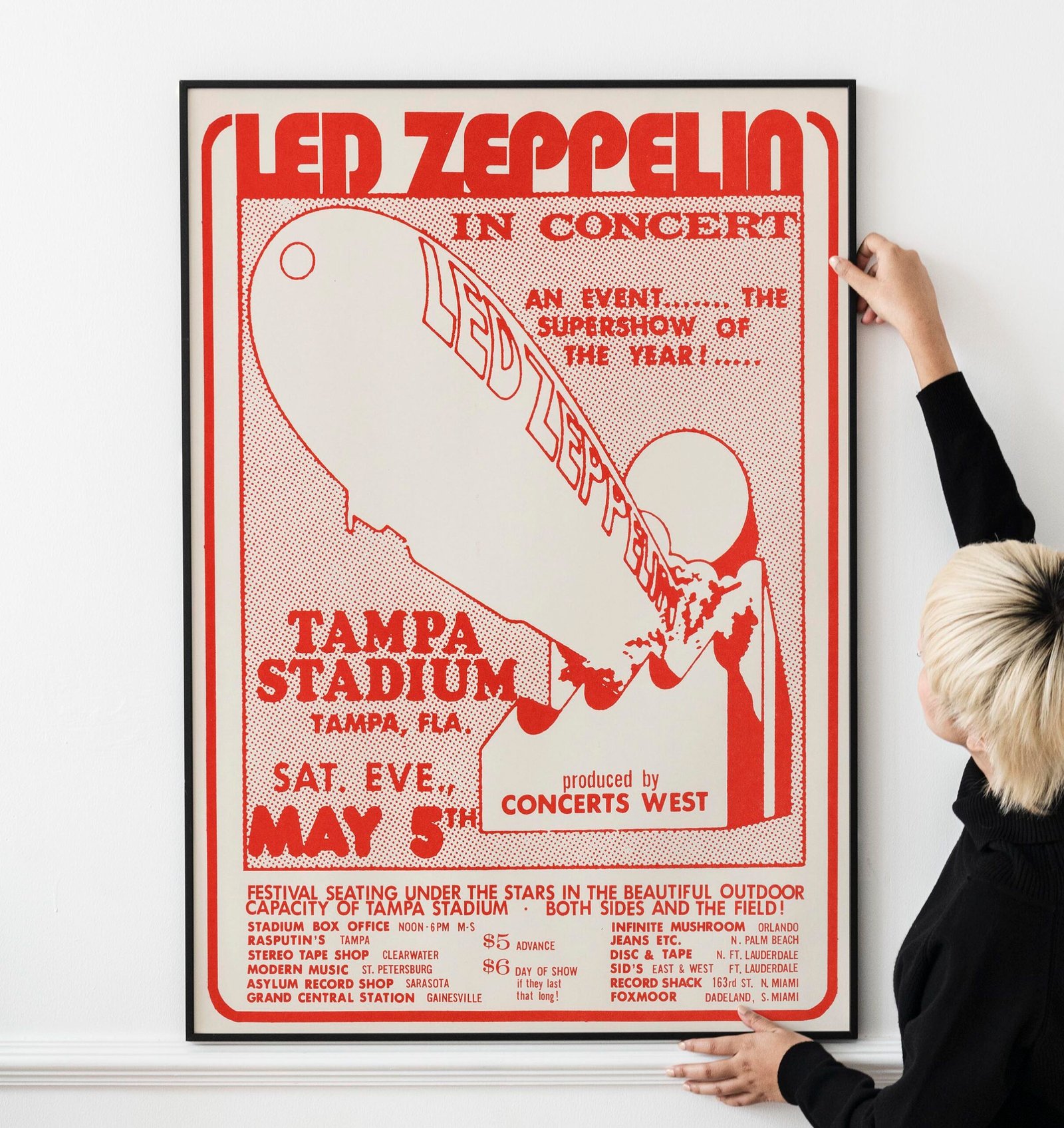 Blond haired Asian woman hanging a frame mockup on a white wall Led Zeppelin 1973 Tampa Concert Poster – Houses of the Holy Tour Print